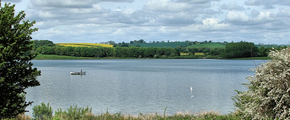 A boat on Rutland Water on a sunny day, with fields in the distance