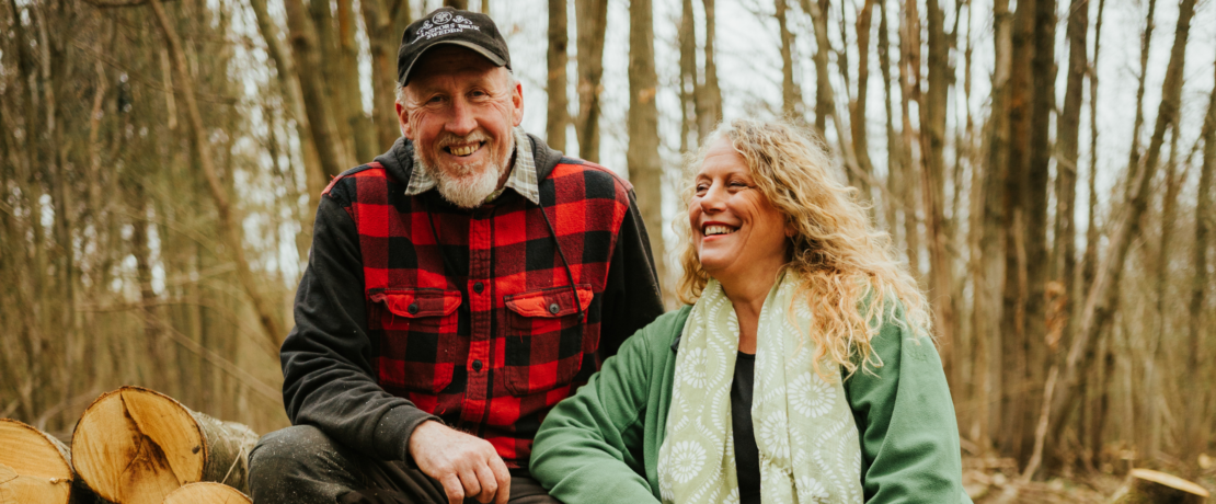 Paul and Lorraine Matthews resting on a log pile in a wooded area