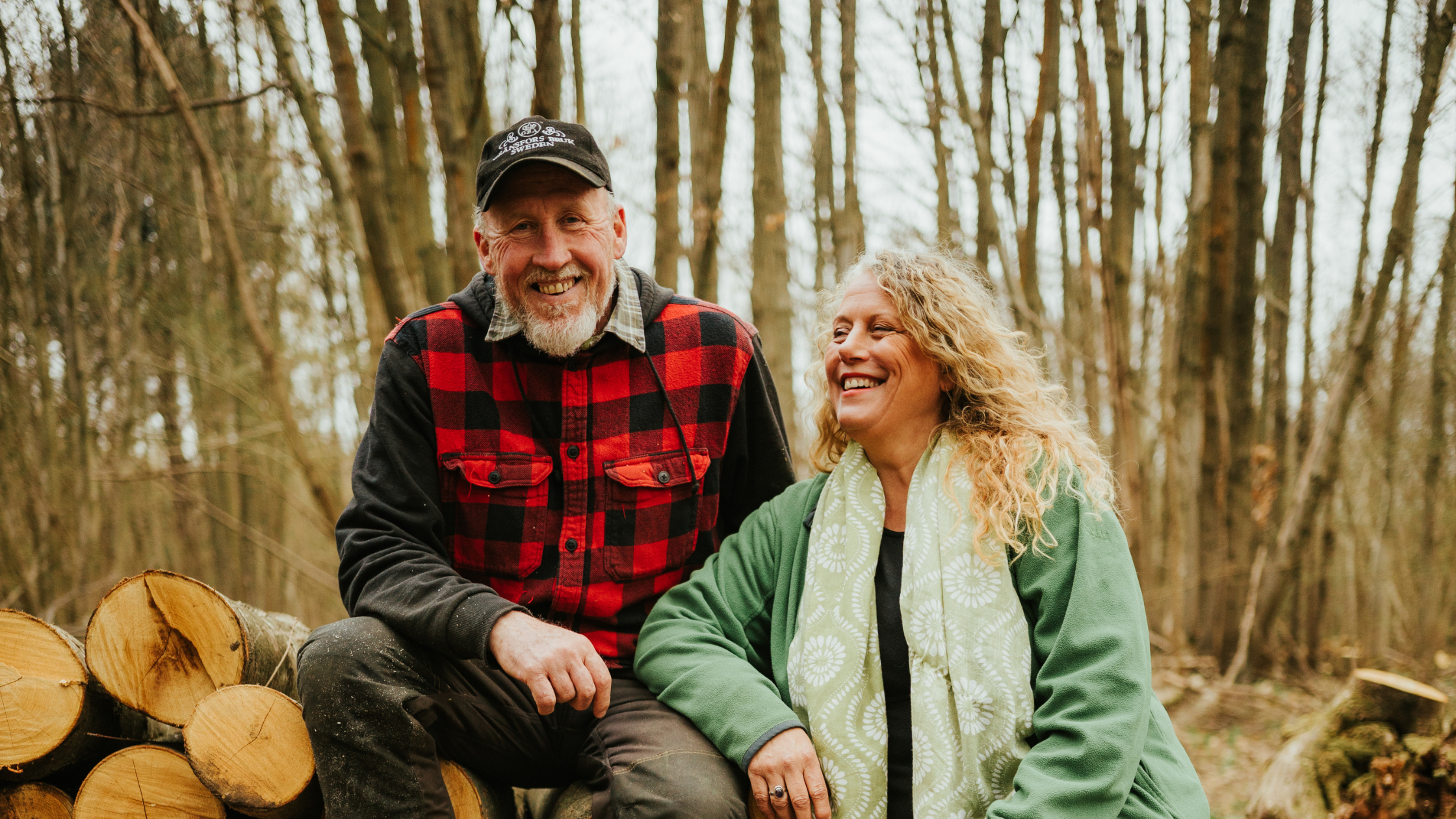 Paul and Lorraine Matthews resting on a log pile in a wooded area