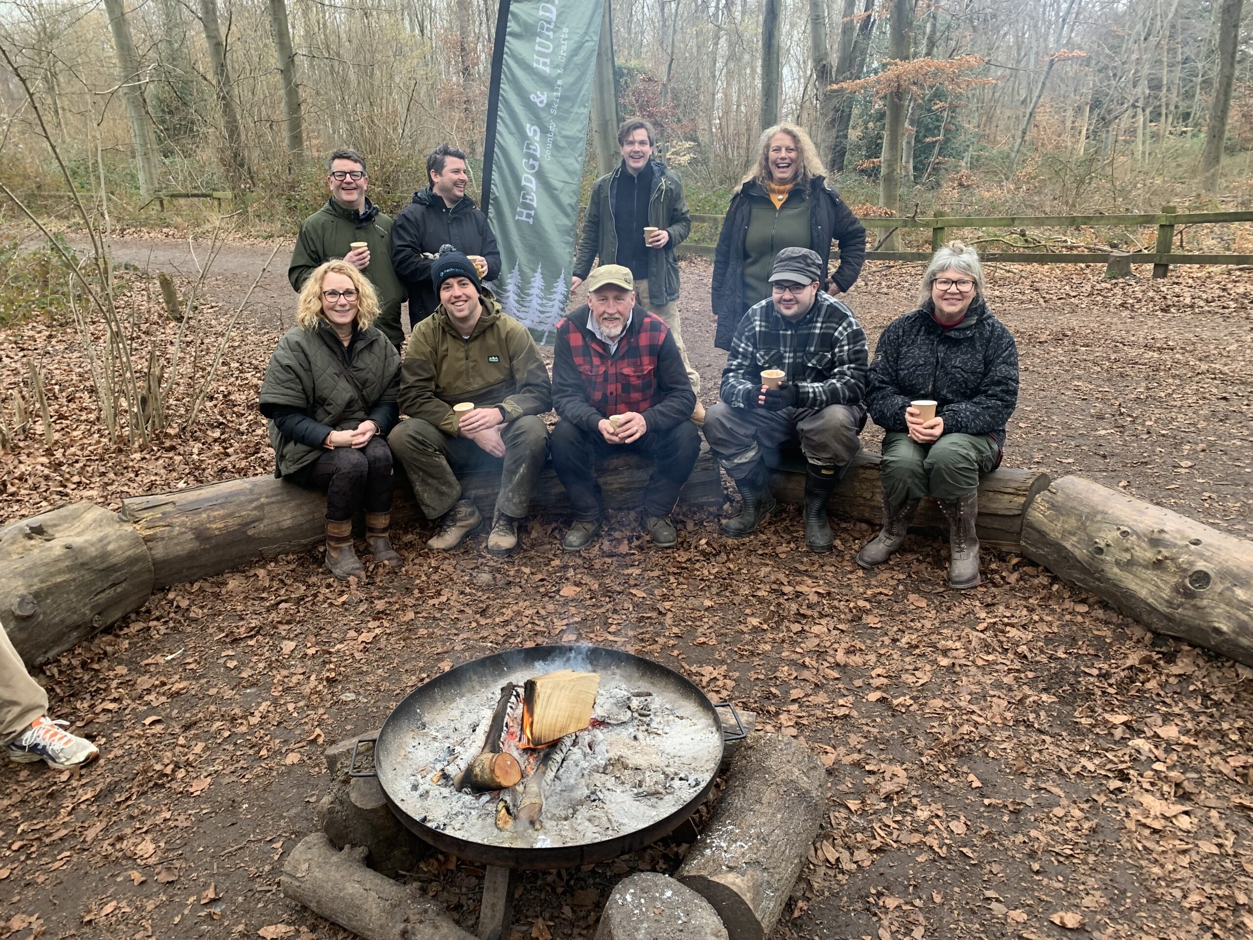 Group of people in a wooded area sat on a log in front of open fire