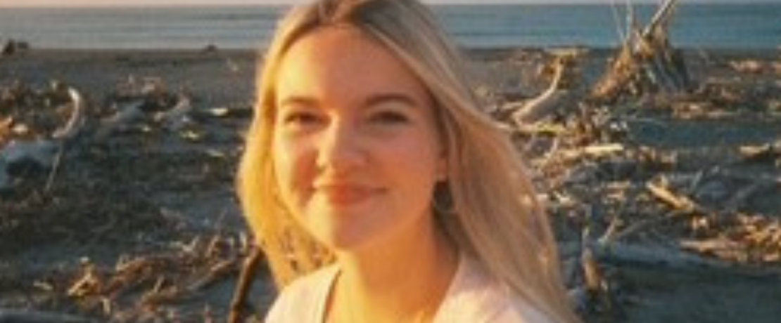 Image of Trustee Emily Johnson. Image shows the head and shoulders of a girl in her early twenties. She is wearing a white cardigan and has blonde hair and is smiling. Int he background is a beach with beach and sea debrie across the sand. You can see the sky disappearing into the horizon with light cloud cover