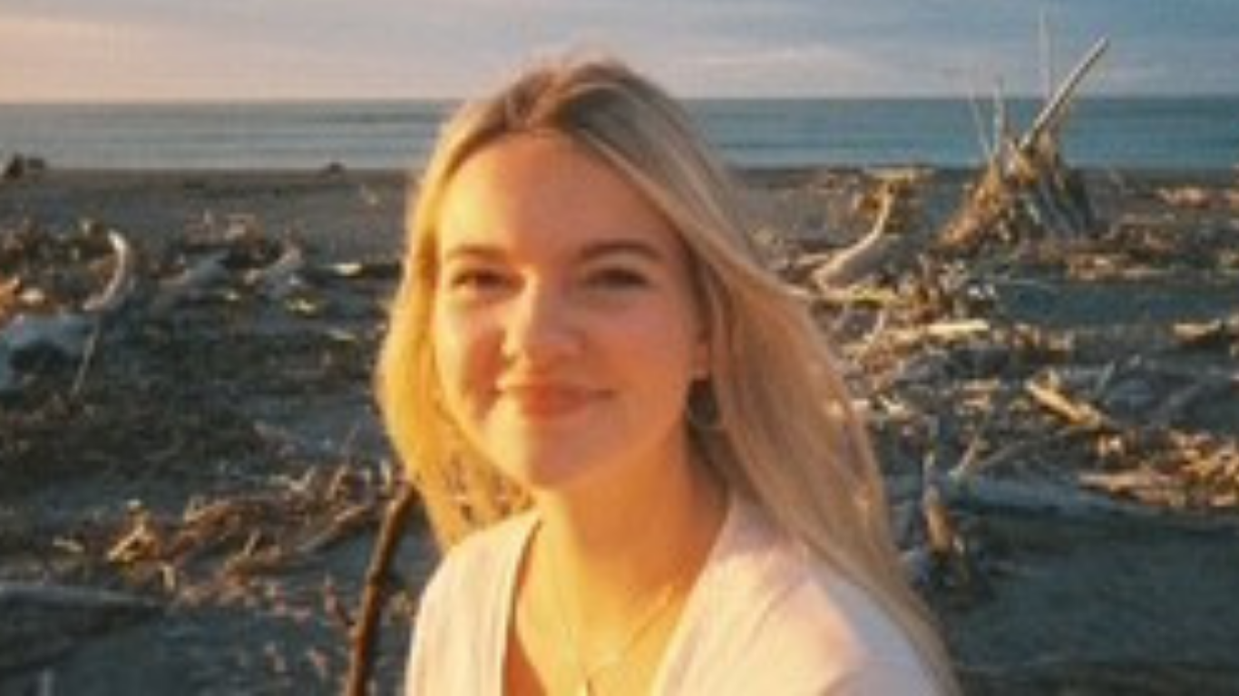 Image of Trustee Emily Johnson. Image shows the head and shoulders of a girl in her early twenties. She is wearing a white cardigan and has blonde hair and is smiling. Int he background is a beach with beach and sea debrie across the sand. You can see the sky disappearing into the horizon with light cloud cover
