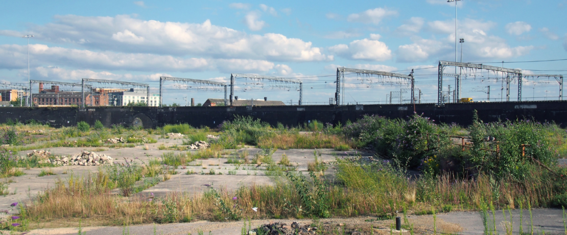 Image of a brownfield site. Image shows a large disused urban site. Covered in cracked concrete with weeds and grass growing through. In the background is a train train and some urban buildings. There is a blue sky with some scattered white clouds.