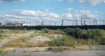 Image of a brownfield site. Image shows a large disused urban site. Covered in cracked concrete with weeds and grass growing through. In the background is a train train and some urban buildings. There is a blue sky with some scattered white clouds.