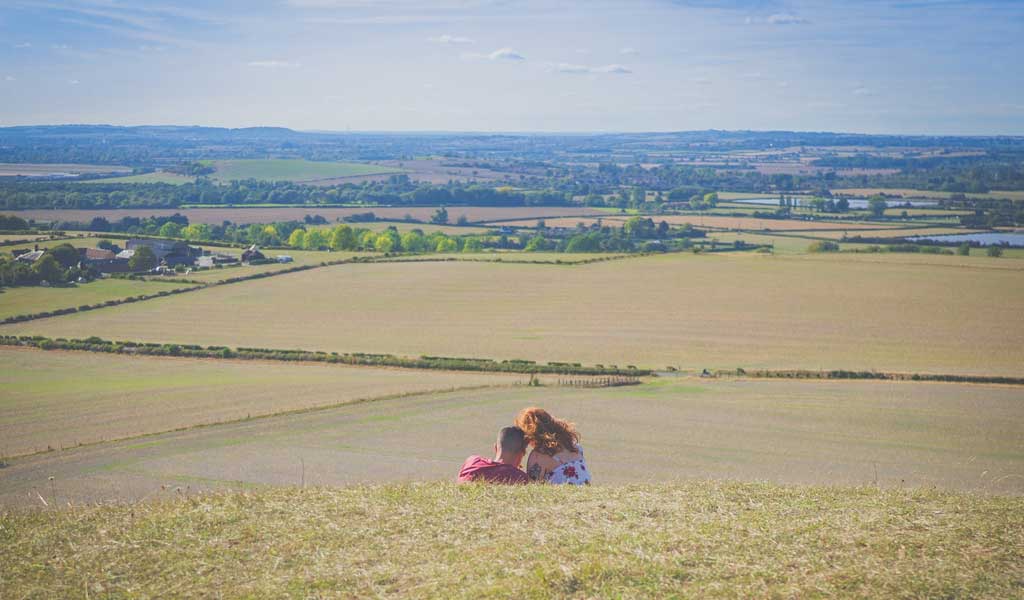 A man and woman lie on grass and lean into each other overlooking a grand vista of fields