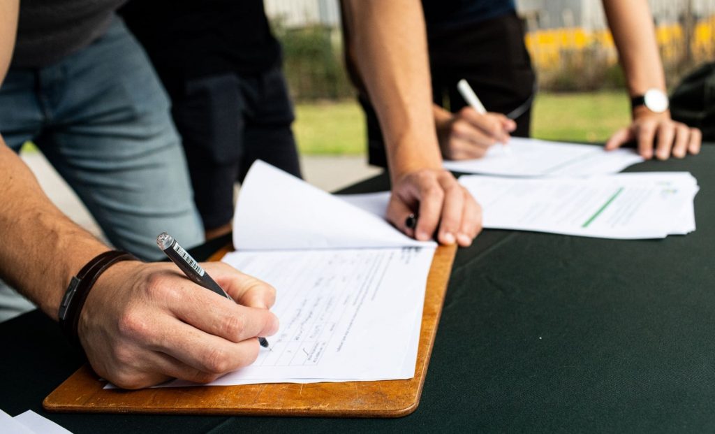 A detail image showing hands signing a piece of paper attached to a clipboard