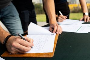 A detail image showing hands signing a piece of paper attached to a clipboard