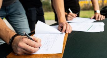 A detail image showing hands signing a piece of paper attached to a clipboard