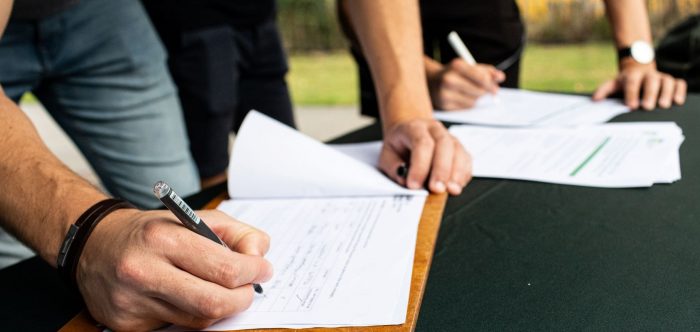 A detail image showing hands signing a piece of paper attached to a clipboard