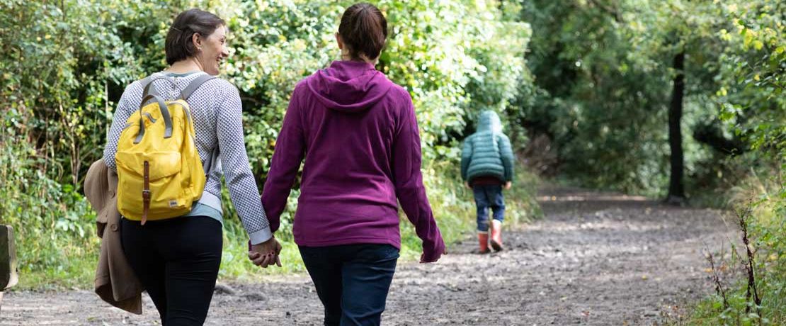 A female couple walk hand in hand through a forest as a small boy runs ahead