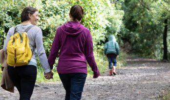 A female couple walk hand in hand through a forest as a small boy runs ahead