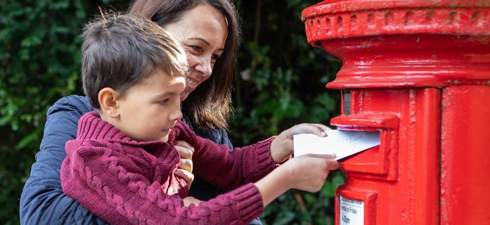 A woman holds a child up so he can post a letter into a red post box