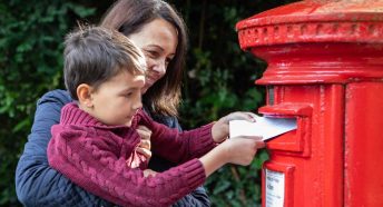 A woman holds a child up so he can post a letter into a red post box