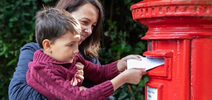 A woman holds a child up so he can post a letter into a red post box