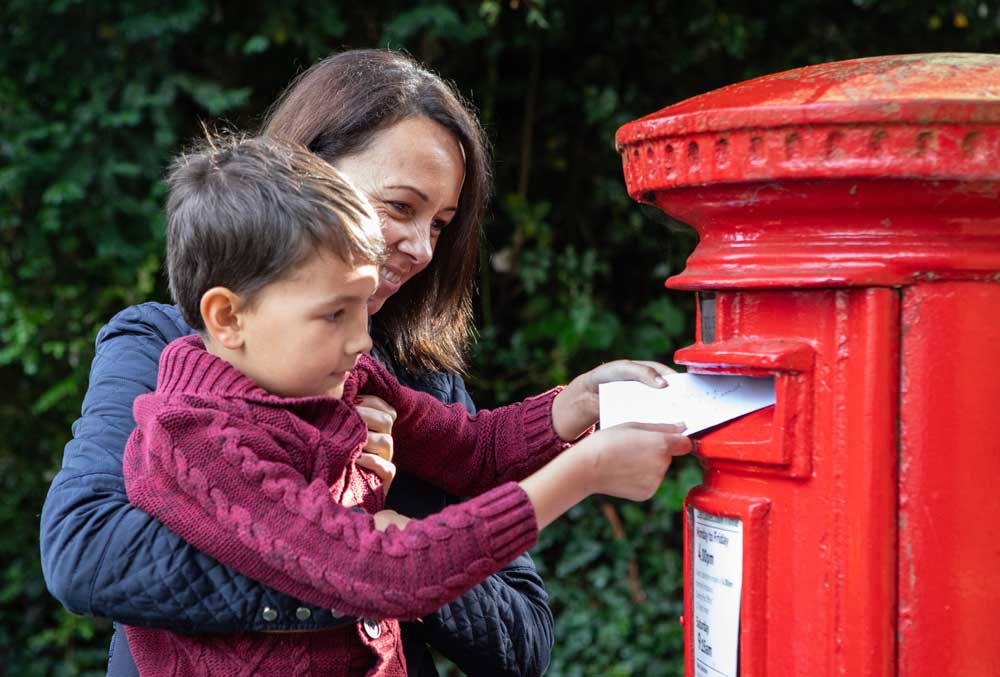 A woman holds a child up so he can post a letter into a red post box