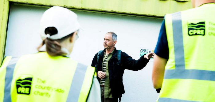 A man stands in front of people in CPRE-branded hugh vis jackets and gives instructions