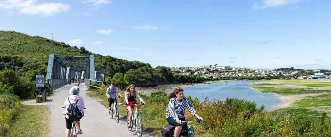 Three young people cycle towards the camera and away from a railway-style bridge over a body of water below blue skies