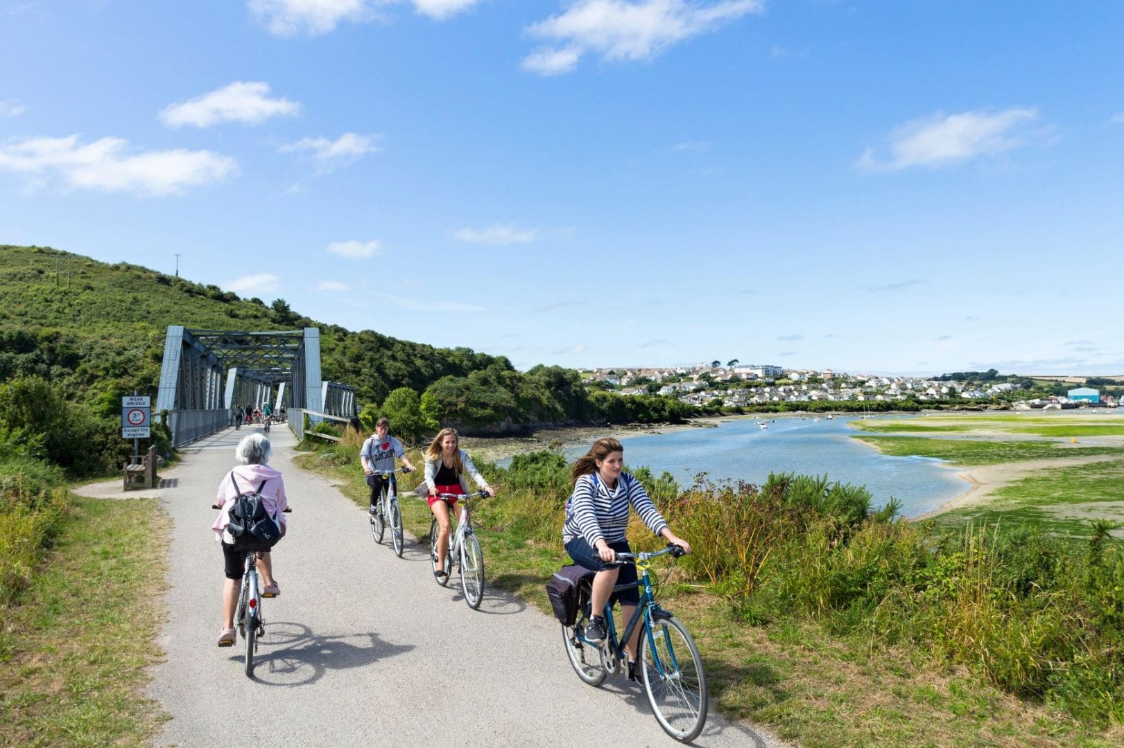 Three young people cycle towards the camera and away from a railway-style bridge over a body of water below blue skies