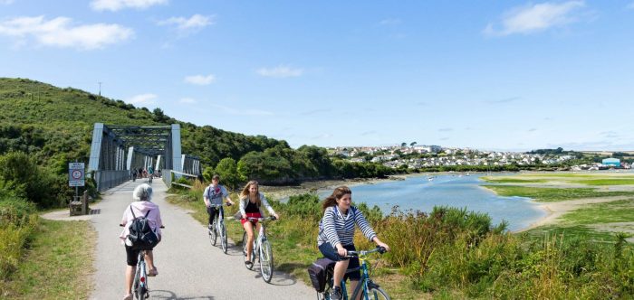 Three young people cycle towards the camera and away from a railway-style bridge over a body of water below blue skies