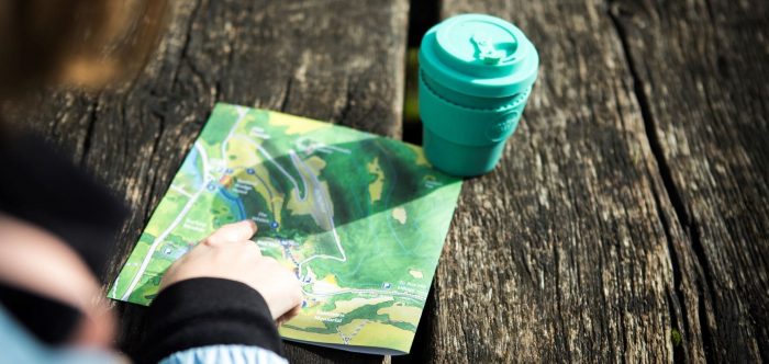 A close-up shot of a map and coffee cup on a picnic table, with someone's finger on the map