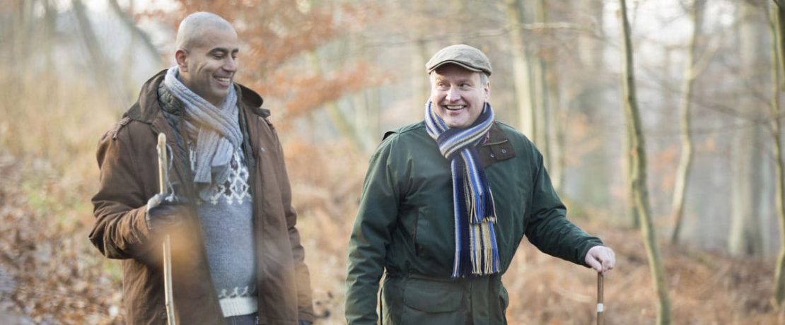 Two men walk through woods with autumn colours behind