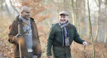 Two men walk through woods with autumn colours behind