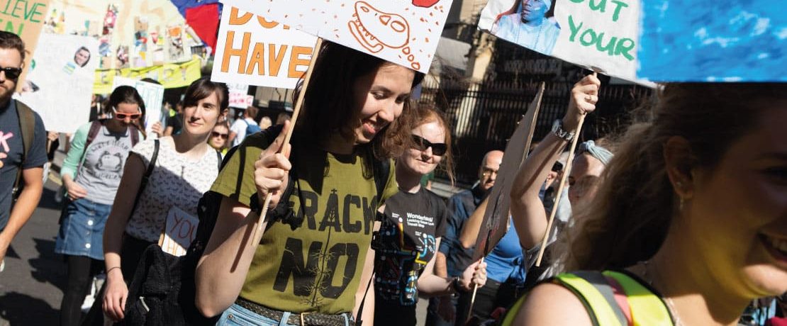 A young woman wearing a 'Frack No' t-shirt holds a banner in a protest march