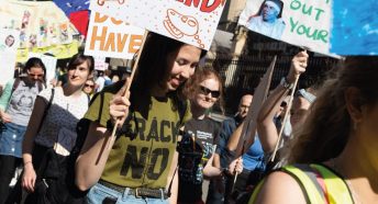 A young woman wearing a 'Frack No' t-shirt holds a banner in a protest march