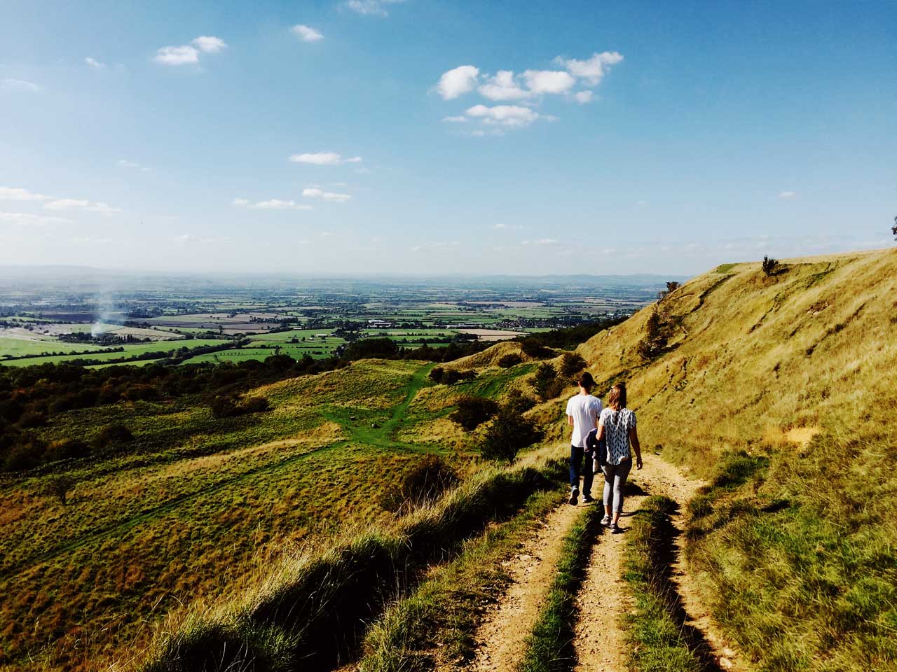 Two people walk along a downhill grassy path with a countryside panorama ahead of them