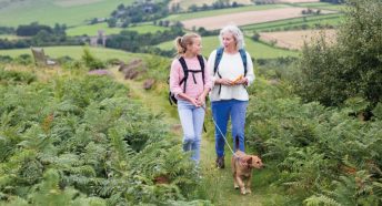 A girl and woman walk a dog through ferns in a green landscape