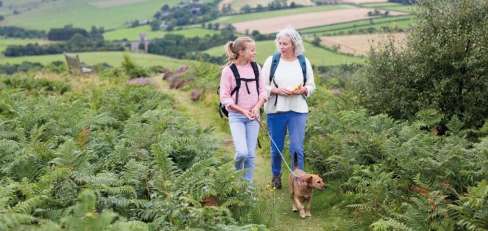 A girl and woman walk a dog through ferns in a green landscape