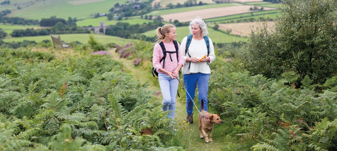 A girl and woman walk a dog through ferns in a green landscape