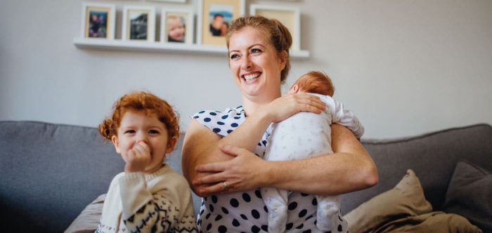 A woman holds a baby and sits beside a child on her sofa