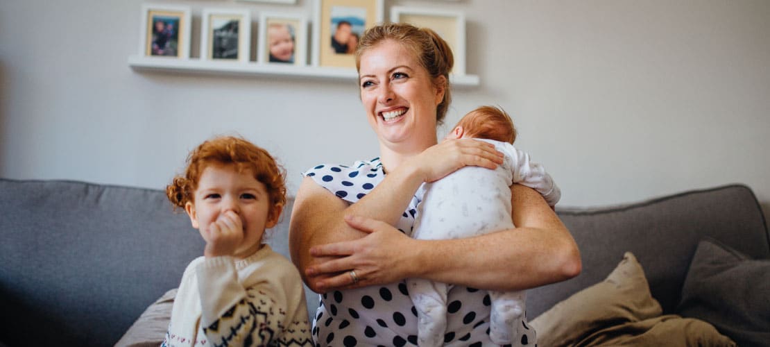 A woman holds a baby and sits beside a child on her sofa