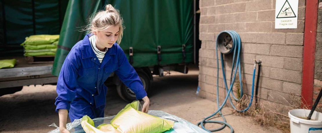 A young woman in blue overalls lifts a large sack on a farm.