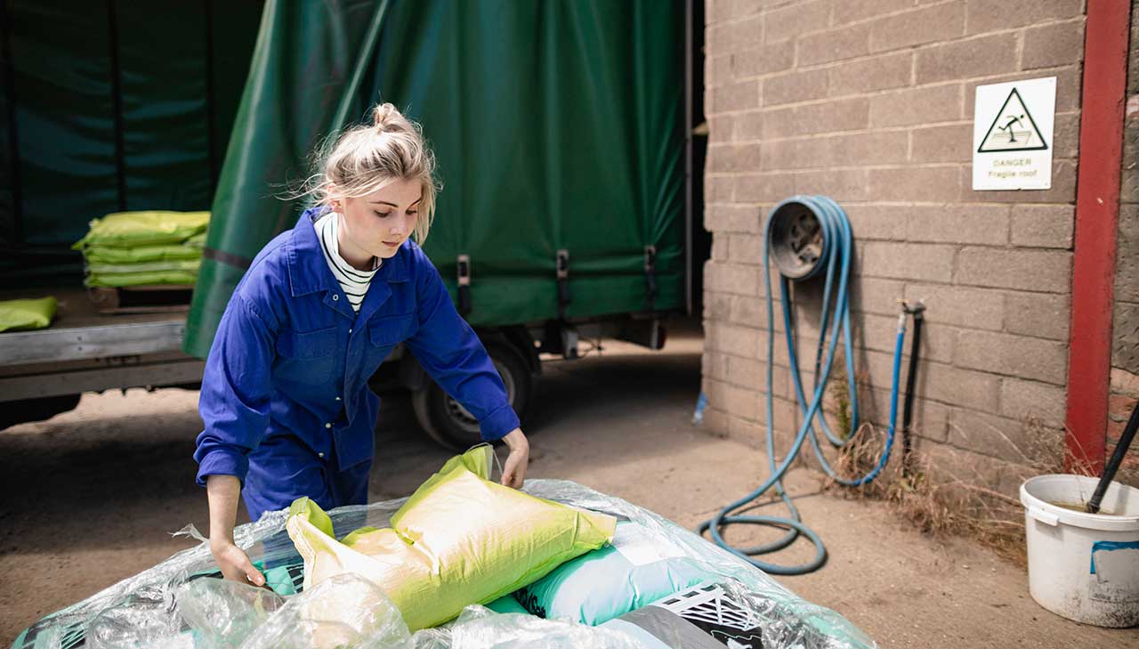 A young woman in blue overalls lifts a large sack on a farm.