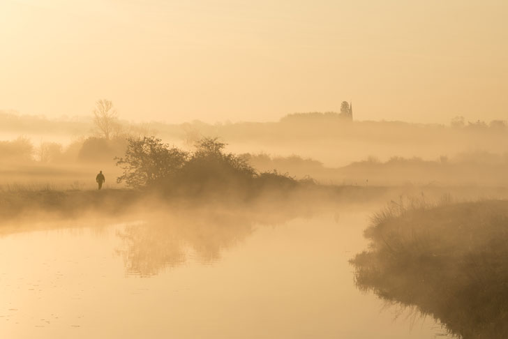 A misty scene across a river with a figure walking along the bank