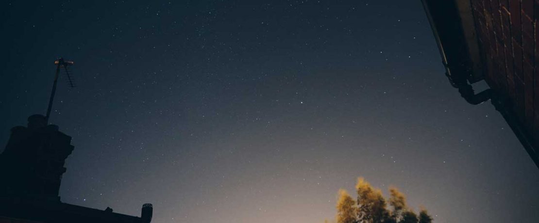 A view of starry skies, with some flare from lights near the horizon, over the roof of houses