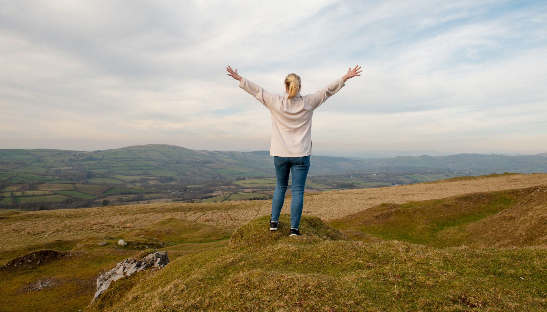 A woman stands at the top of a hill with arms outstretched above her head