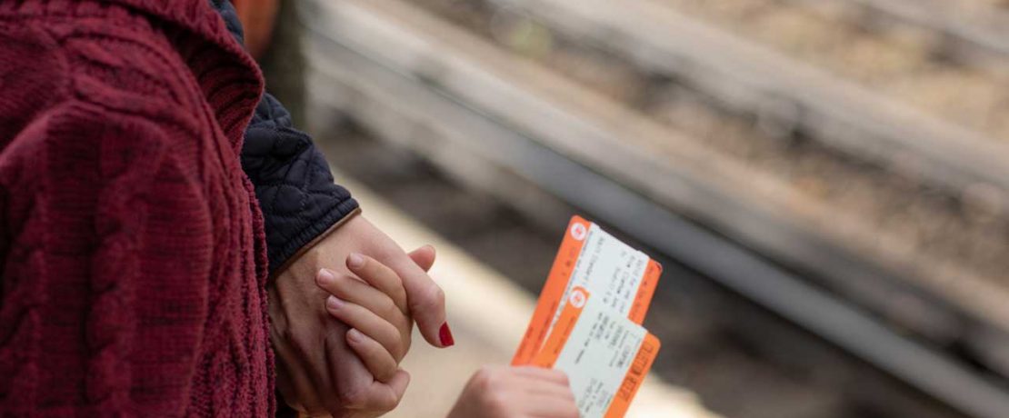 A detail of a woman holding a child's hand; the child holds train tickets