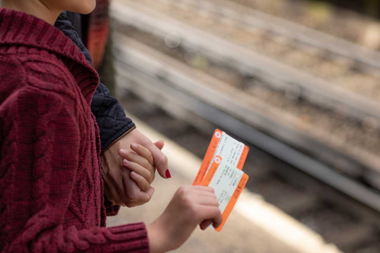A detail of a woman holding a child's hand; the child holds train tickets