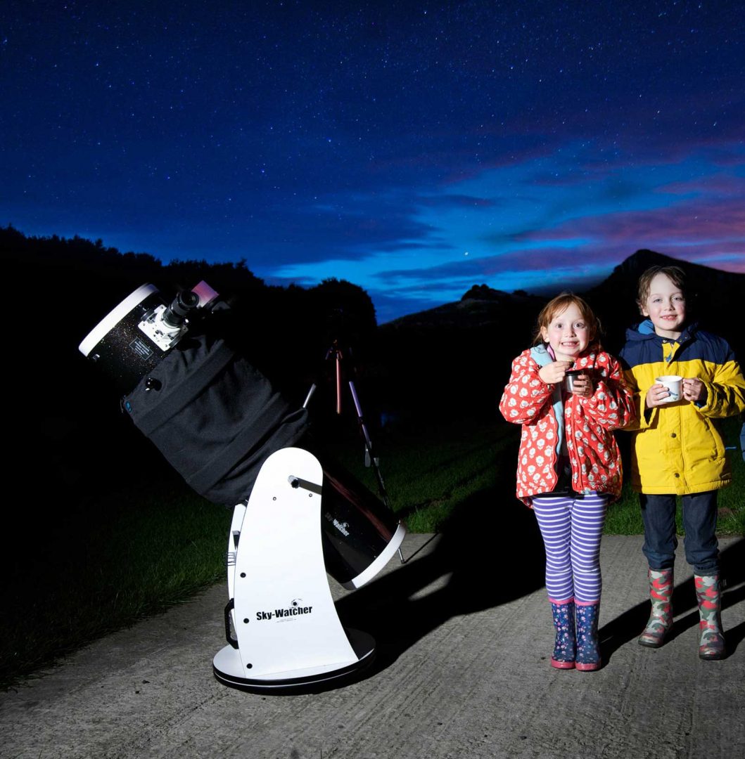 Two girls stand beside a large telescope with a dark night sky behind them
