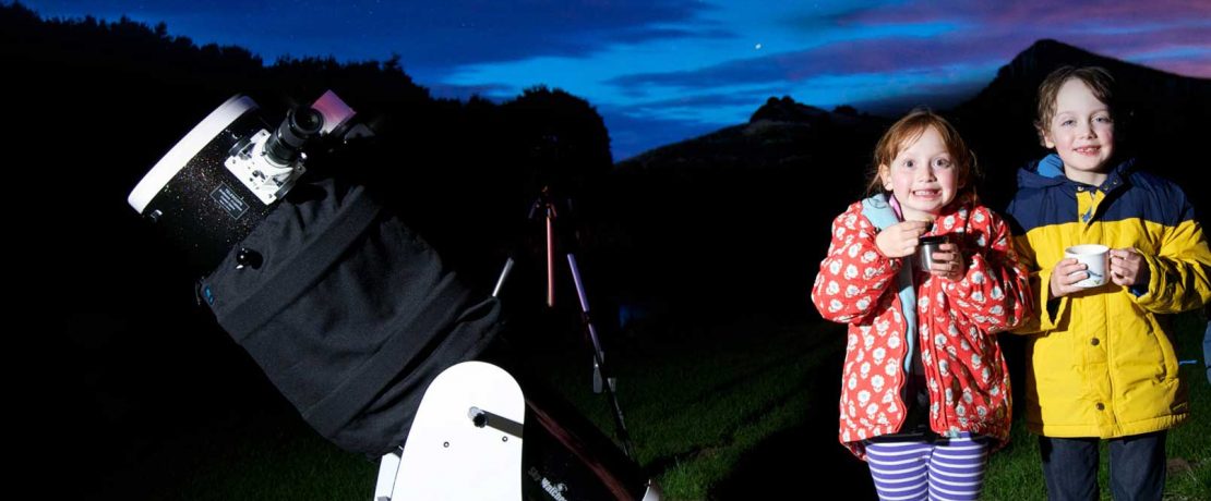 Two girls stand beside a large telescope with a dark night sky behind them