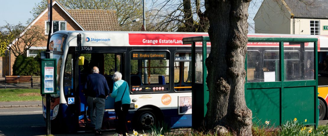 Two elderly people are seen from behind as they climb onto a bus in a rural setting