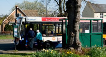 Two elderly people are seen from behind as they climb onto a bus in a rural setting