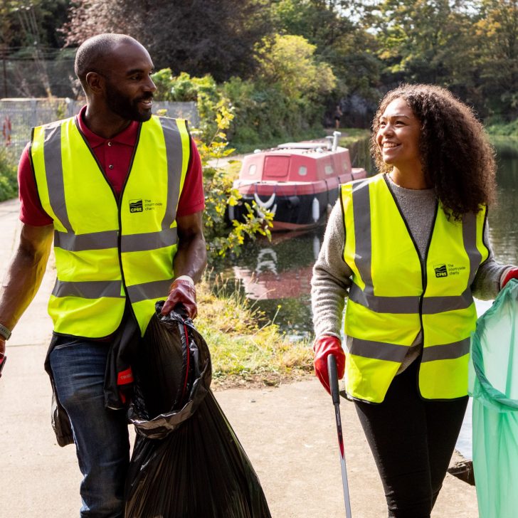 A man and a woman look at one another as they walk with bin bags and litter collecting tools beside a canal