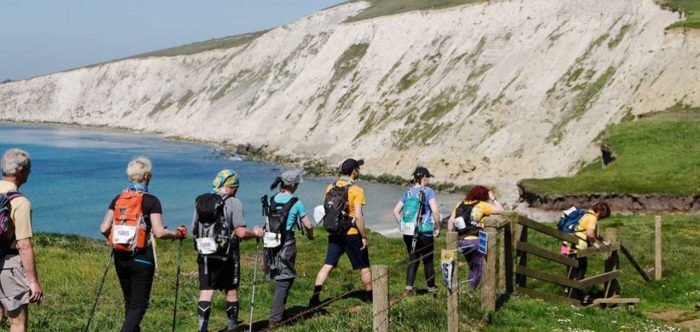 A train of people wearing challenge numbers on rucksacks walk along the top of white cliffs