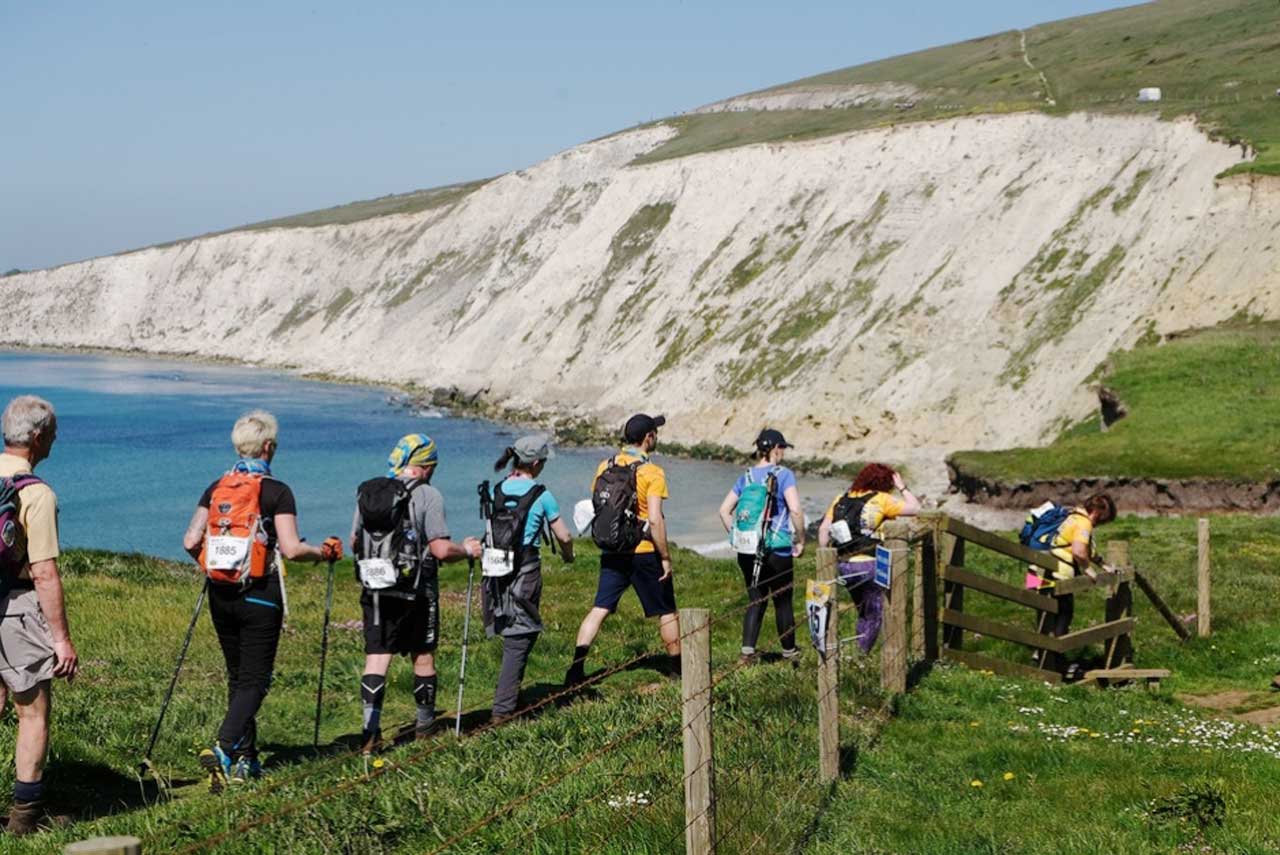 A train of people wearing challenge numbers on rucksacks walk along the top of white cliffs