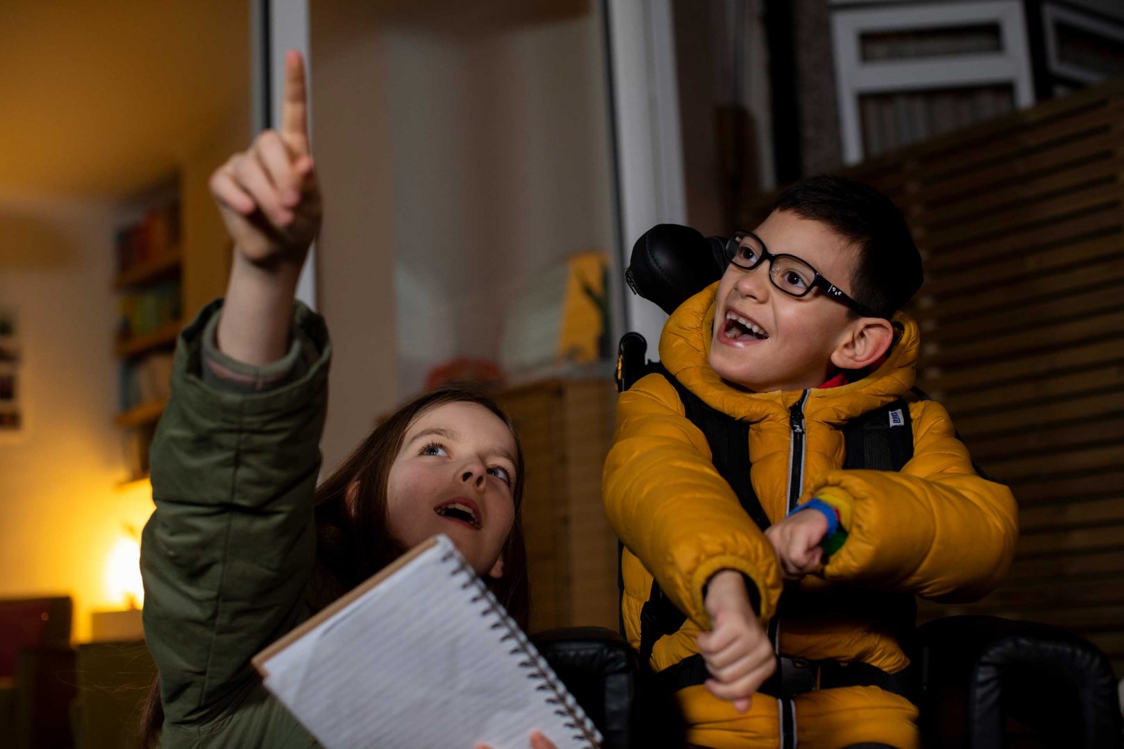 A boy in a wheelchair wearing a bright jacket looks up as a young girl points to the night sky