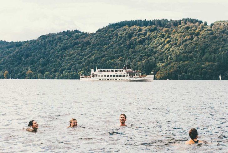 Four people's heads are visible above pale water with a lake and wooded slope behind